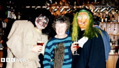 A group of three people standing inside a traditional pub with a wooden bar and shelves of glasses in the background. The individuals are dressed in costumes: one in a tattered outfit with boots holding a pint of beer, another in a striped sweater holding a broomstick, and the third in a long dark robe with green hair-like material, holding a glass of wine.