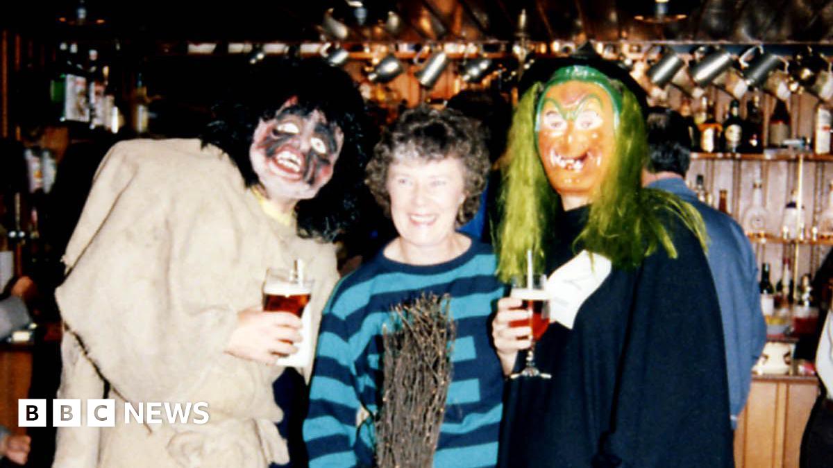 A group of three people standing inside a traditional pub with a wooden bar and shelves of glasses in the background. The individuals are dressed in costumes: one in a tattered outfit with boots holding a pint of beer, another in a striped sweater holding a broomstick, and the third in a long dark robe with green hair-like material, holding a glass of wine.