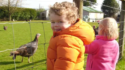 Verity and Ian A young boy with curly blond hair wearing a bright orange padded jacket turns and smiles cheekily at the camera, while next to him is a fence and some turkeys strutting behind it. A younger girl with straight blond hair wearing a pink fleece stands next to him, with her face turned away from the camera.