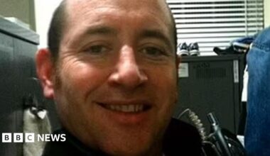 A selfie taken by David Carrick of him sitting in an office with a filing cabinet behind him. He is smiling and has a walkie-talkie strapped to his chest.