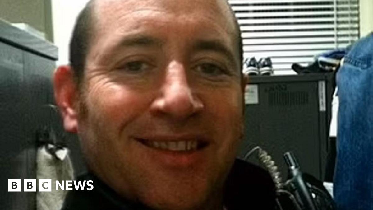 A selfie taken by David Carrick of him sitting in an office with a filing cabinet behind him. He is smiling and has a walkie-talkie strapped to his chest.