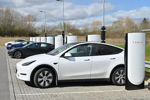 John Keeble/Getty Images A white Tesla Electric EV car charges at a Tesla Public Supercharger network, charging station