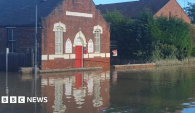 A brick building with a red door and arched windows outlined in white face on to a road which is underwater.
