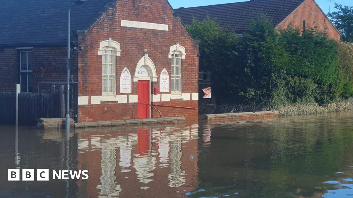 A brick building with a red door and arched windows outlined in white face on to a road which is underwater.