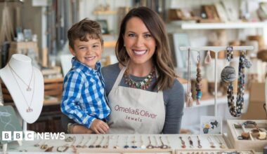 An AI-generated image showing a woman with brown hair holding a child in her arms and standing next to a table filled with necklaces.