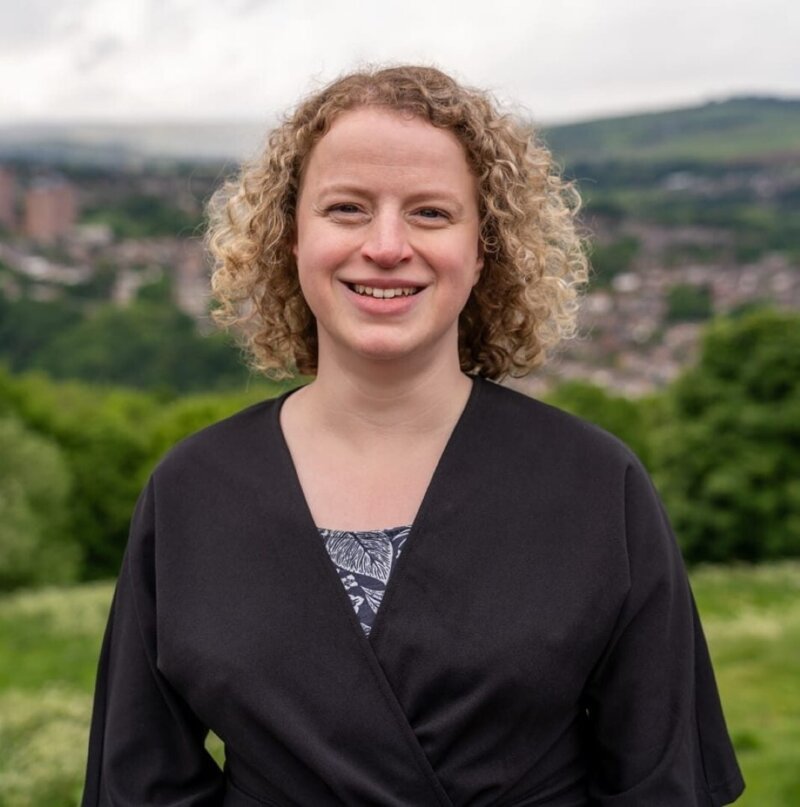 A profile shot of a woman with medium length curly blond hair smiling directly at the camera in an outdoor setting