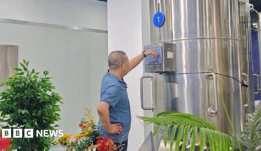 A man in a blue t-shirt stands looking at a large metallic container which has the letters YFCYRO on it. Around him are several green plants.