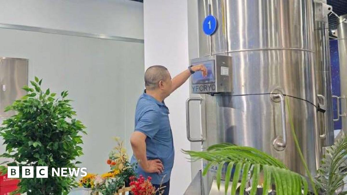 A man in a blue t-shirt stands looking at a large metallic container which has the letters YFCYRO on it. Around him are several green plants.