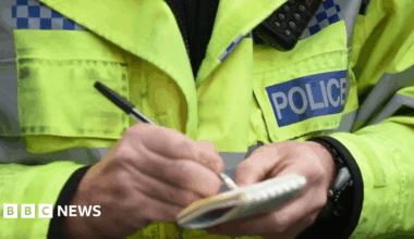 A police officer wearing a yellow police jacket and holding a note pad and pen.