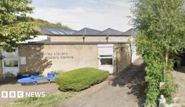 A single-storey, cream-coloured stone building, with a paved area to the front where there is a blue 'S'-shaped bench.