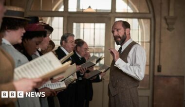 A group of people in historical costume stand in a line, holding sheet music, mid-rehearsal. A man, Ralph Fiennes, in a white shirt and waistcoat gestures with one hand, likely conducting.