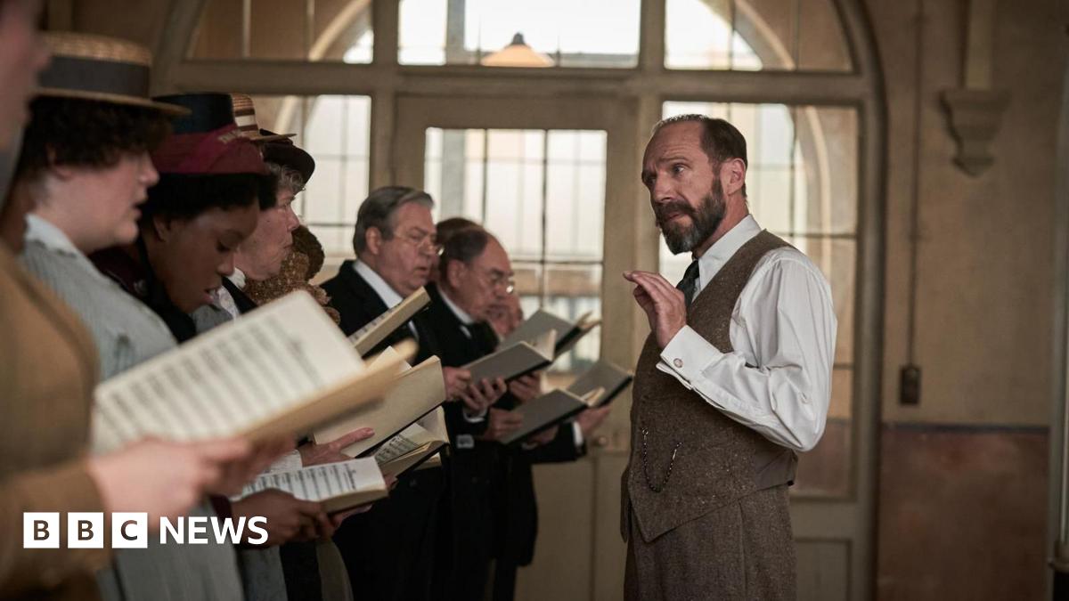 A group of people in historical costume stand in a line, holding sheet music, mid-rehearsal. A man, Ralph Fiennes, in a white shirt and waistcoat gestures with one hand, likely conducting.