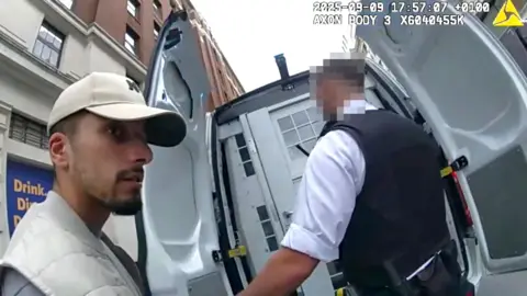 Metropolitan Police Brahim Kaddour-Cherif seen in arrest footage from police bodycam. He is wearing a baseball cap and white sleeveless coat with a grey top on underneath. He is being led into a police van by an officer wearing body armour.
