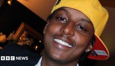 A man wearing a sideways yellow cap and a silver earring in his left ear is smiling at the camera. The image is a close-up so only his head is visible. There appears to be a photo frame in the background but it is blurry.