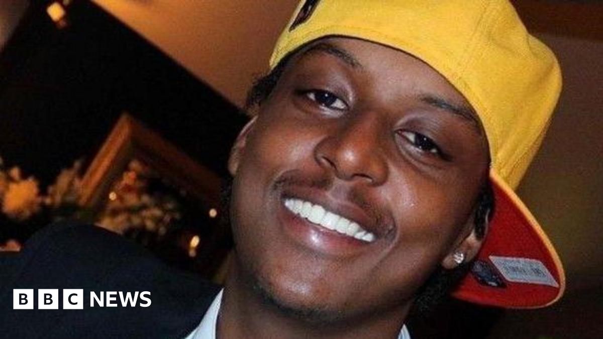 A man wearing a sideways yellow cap and a silver earring in his left ear is smiling at the camera. The image is a close-up so only his head is visible. There appears to be a photo frame in the background but it is blurry.