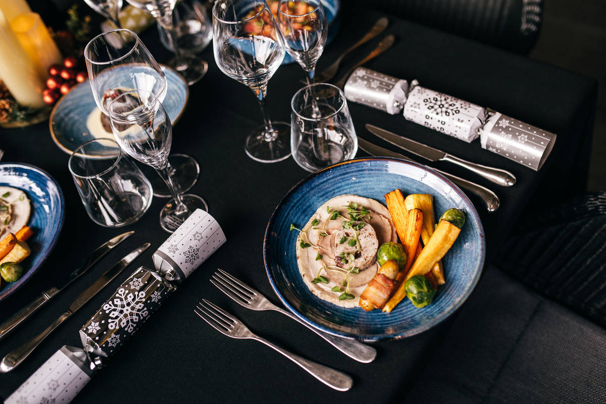 a festive place setting at a dark panelled table in a londonr restaurant, with christmas crackers, and plats of food and glasses of wine