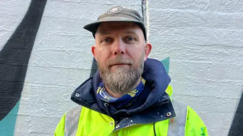 BBC/Chloe Aslett A man with a brown cap, brown and grey beard, and large neon jacket looks at the camera with a neutral expression. The painted white, black and blue wall is just behind him.
