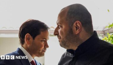 A man in blue suit, white shirt and red tie stands close to a man in a dark jacket.