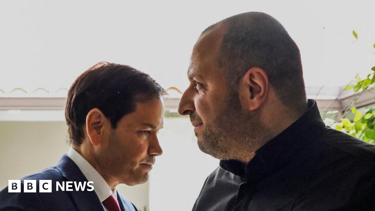 A man in blue suit, white shirt and red tie stands close to a man in a dark jacket.