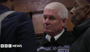 Paul Costelloe has receding grey hair. He is wearing a bright pink, blue and green scarf and a blue jacket.