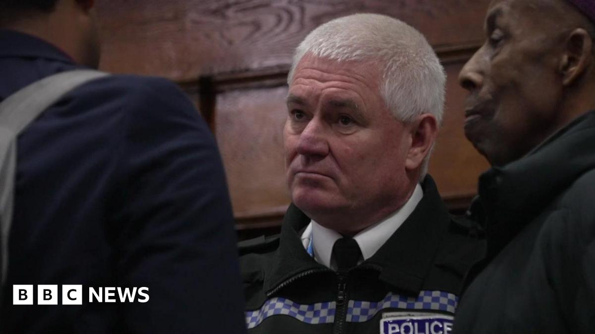 Paul Costelloe has receding grey hair. He is wearing a bright pink, blue and green scarf and a blue jacket.