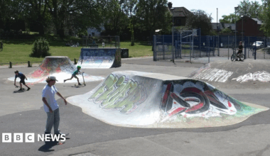 A skate park with two boys using skateboards and another on a scooter. There are skate ramps which have graffiti tags on them. In the background people are sat on some grass under trees.