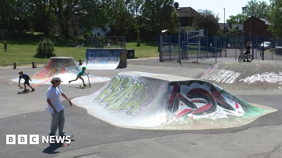 A skate park with two boys using skateboards and another on a scooter. There are skate ramps which have graffiti tags on them. In the background people are sat on some grass under trees.