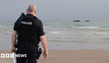 A French police officer stands on the beach at Gravelines as a French maritime police rib approaches a small boat off the coast in the background, taken in July.