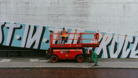Danni Maibaum A man wearing a high-visibility orange vest stands on a red mobile elevated work platform in front of a large tiled wall. The wall features a bold mural with large black and white letters on a light blue background. The platform is positioned on a pavement next to a green safety barrier.
