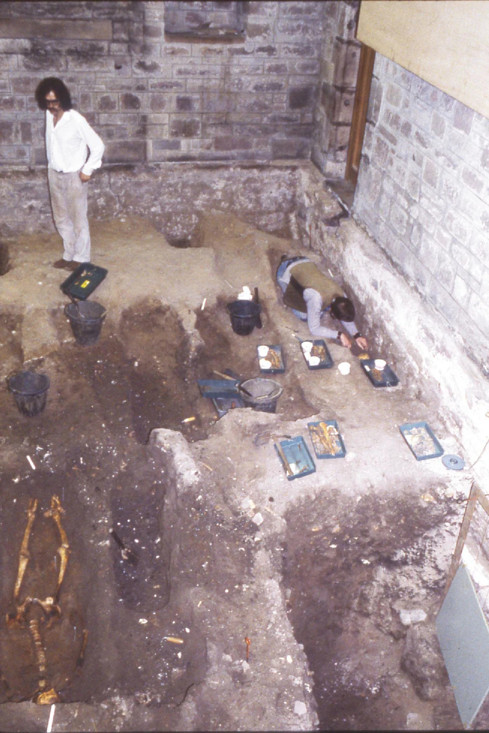 One archaeologist is standing beside open graves. He is wearing a white shirt and has shoulder-length brown hair. There is a skeleton in one of the graves. There is another archaeologist lying down while working one another skeleton.