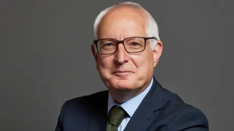 PA Media Neil Duncan-Jordan who is looking directly at the camera and smiling. He is wearing a navy suit jacket with a pale blue shirt and green tie. 