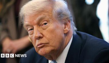 A close up image of Trump in the Oval Office. He wears a dark suit and blue tie