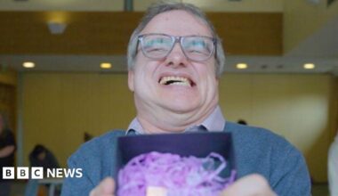 A man smiling broadly, baring his teeth. He is holding a trophy inside a box with purple shredded tissue. He is wearing a blue jumper and a checked shirt.