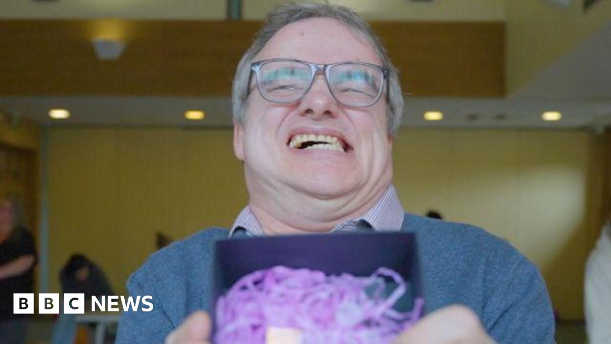 A man smiling broadly, baring his teeth. He is holding a trophy inside a box with purple shredded tissue. He is wearing a blue jumper and a checked shirt.