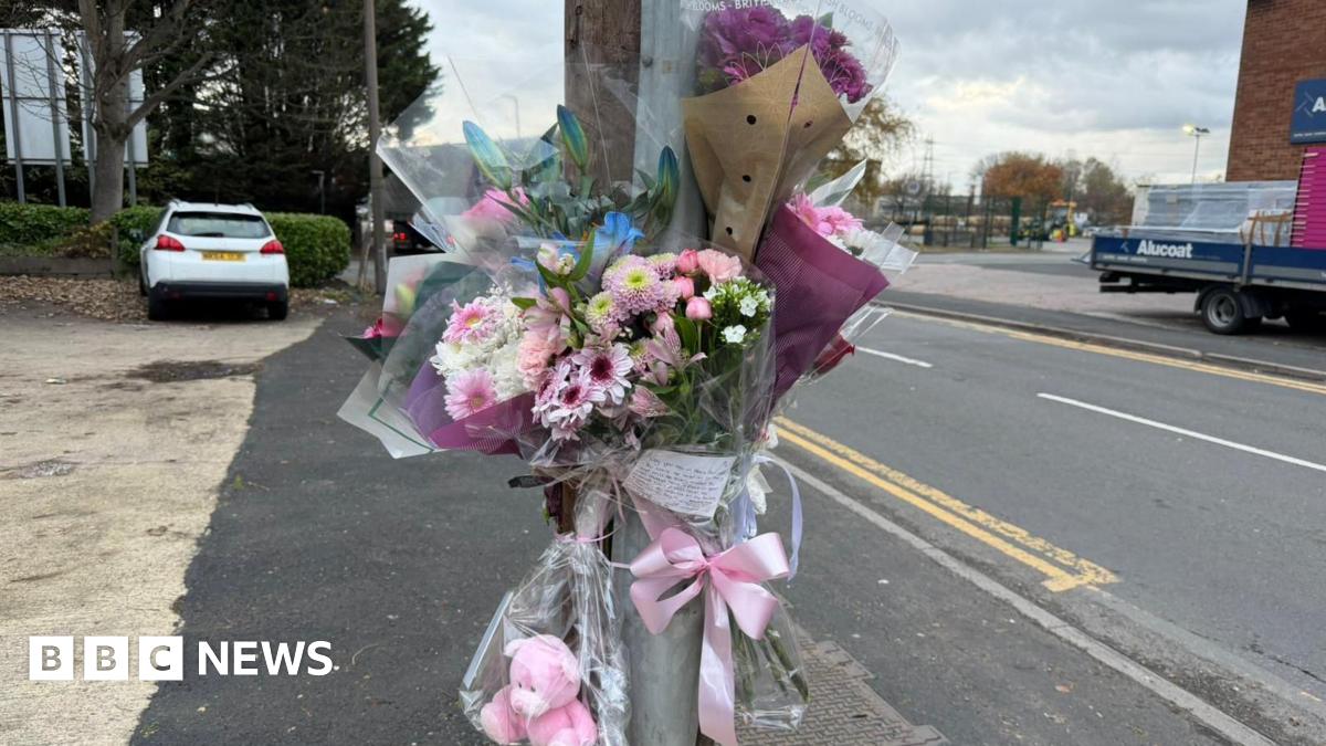 Six bouquets of flowers are tied to a pole with a pink ribbon, a handwritten note and a pink teddy bear, by the side of a road. Vehicles can be seen in the background