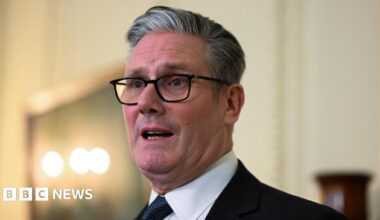 Britain's Prime Minister Keir Starmer looks off camera, wearing his customary black framed glasses and a dark patterned tie.