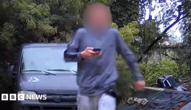 A surveillance photo of a male teenager wearing grey sweat pants and shirt and with his face blurred, carrying a mobile phone. He is walking away from a grey van parked on hard surface in a wooded area.