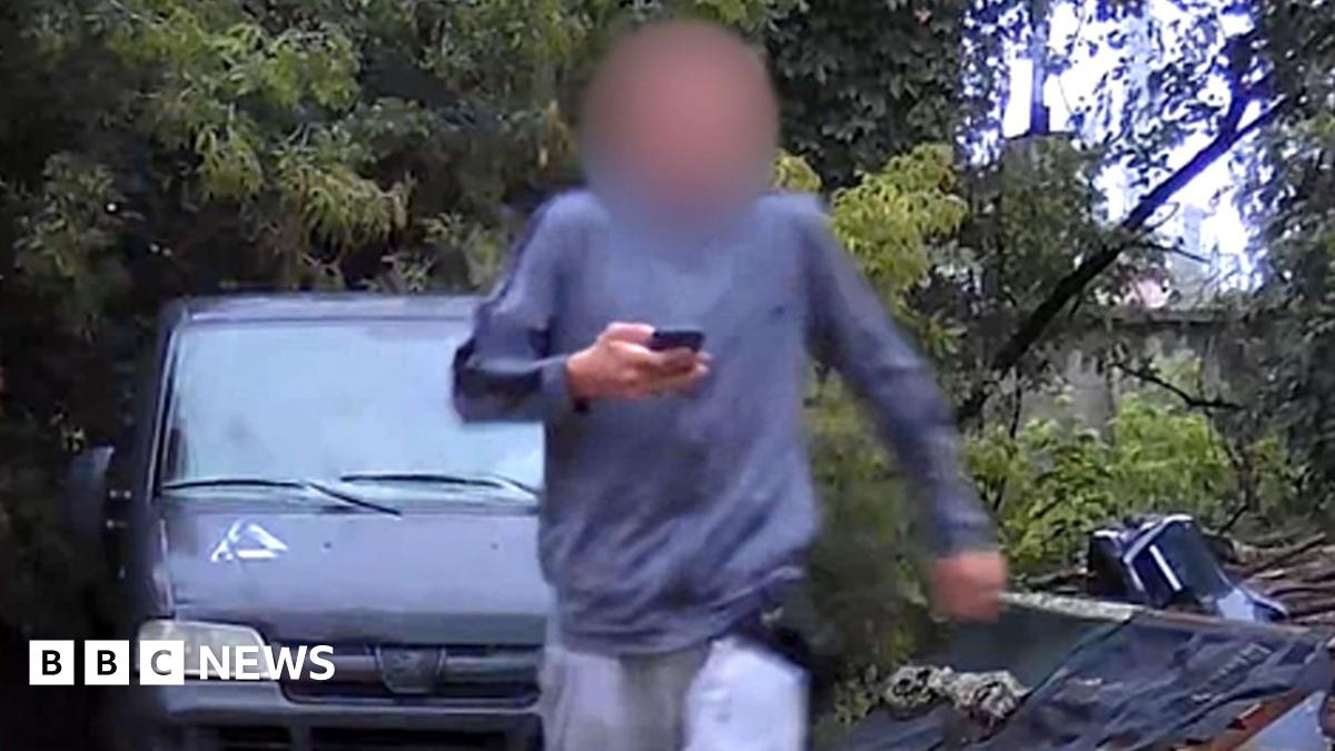 A surveillance photo of a male teenager wearing grey sweat pants and shirt and with his face blurred, carrying a mobile phone. He is walking away from a grey van parked on hard surface in a wooded area.