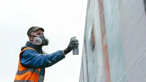 Danni Maibaum A man wearing a blue jacket, orange safety vest, and gloves is using a spray paint can to paint a large vertical surface. He is also wearing a respirator mask, and the wall being painted appears to have light-coloured sections with some geometric shapes.
