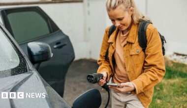 A young woman with her phone in her hand, about to plug an electrical charger into her car - she is wearing an orange jacket and has her blonde hair tied back into a pony tail, and is standing next to the car with the door opened