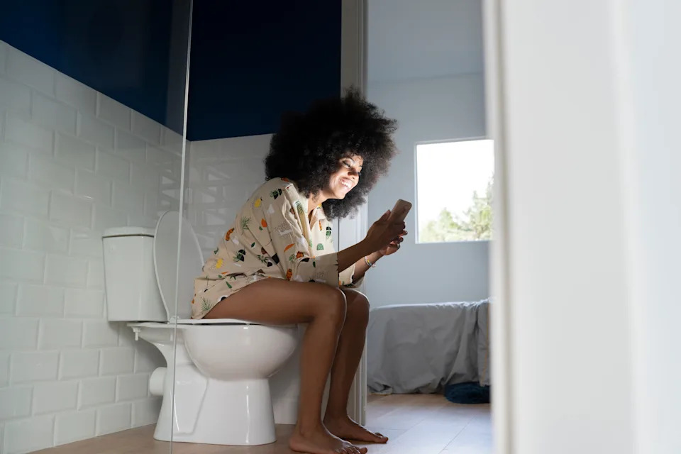 Person sitting on a toilet in a bathroom, smiling at their phone, wearing a light-colored shirt with patterns. Relaxed and casual setting