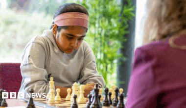 Bodhana wearing a grey sweater, and with a pink headband, stares with deep concentration at a chess set. She is playing against an adult woman.