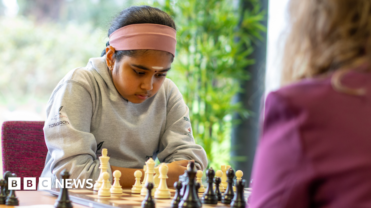 Bodhana wearing a grey sweater, and with a pink headband, stares with deep concentration at a chess set. She is playing against an adult woman.