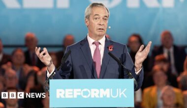 Nigel Farage in a suit and tie standing behind a podium with the words ‘REFORM UK’ printed on the front, speaking into two microphones. The background shows a large blue banner with partial text and an audience seated behind.