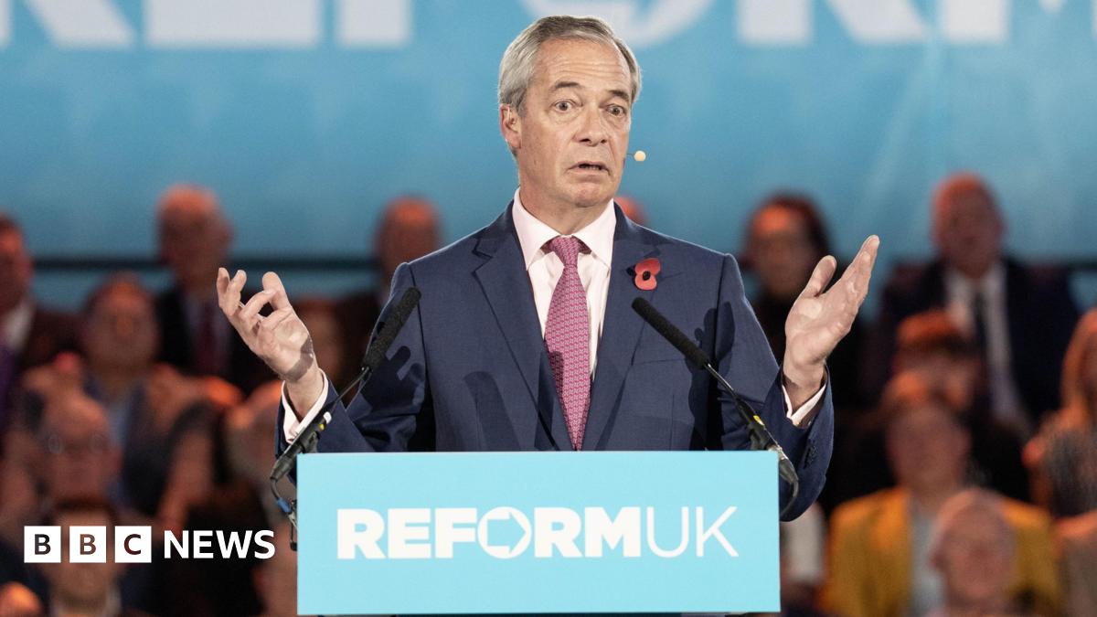 Nigel Farage in a suit and tie standing behind a podium with the words ‘REFORM UK’ printed on the front, speaking into two microphones. The background shows a large blue banner with partial text and an audience seated behind.