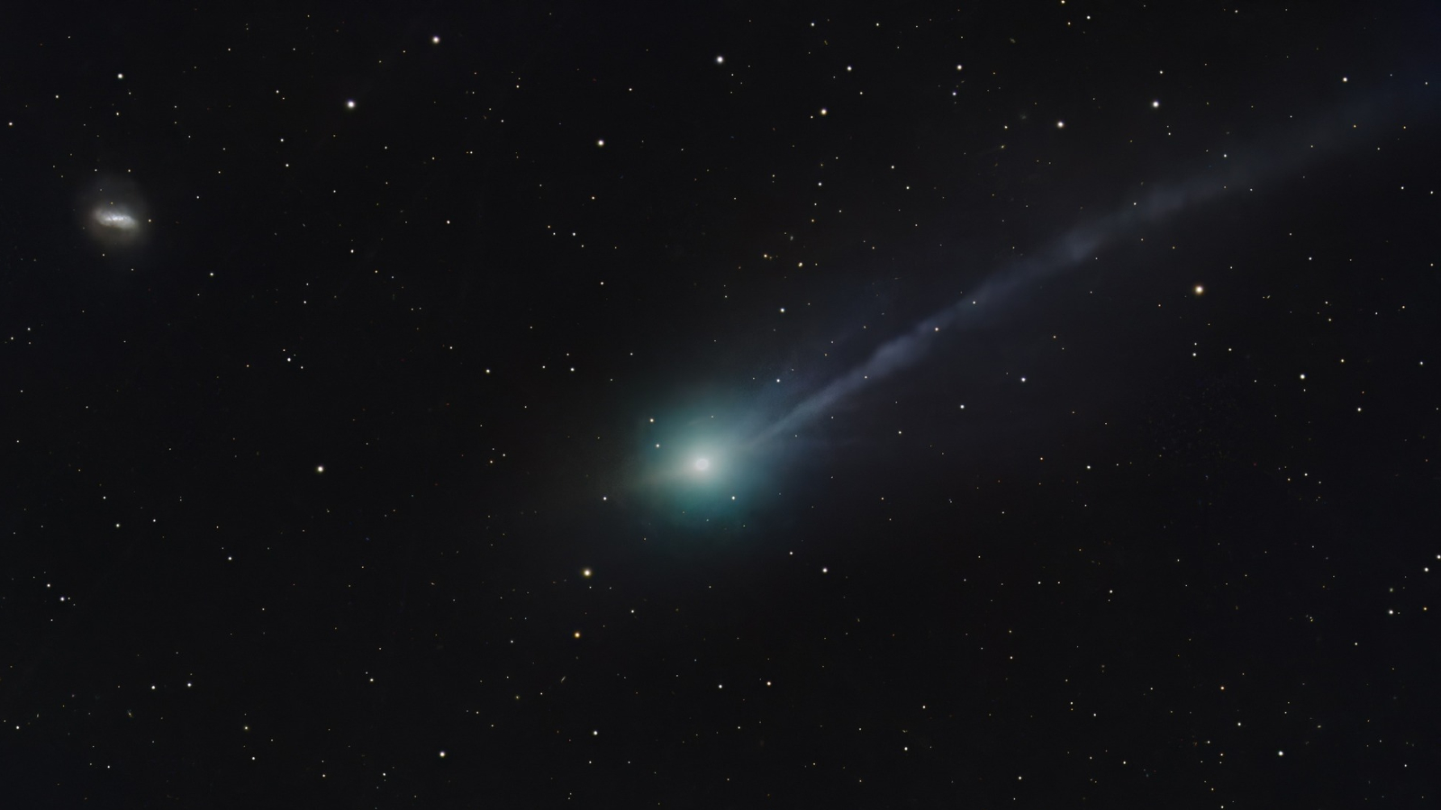 A photo of 3I/ATLAS with a green coma and a long tail, as well as a second shorter tail. A spiral galaxy is also visible in the top left of the image.