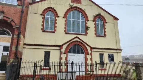 LDRS Pisgah Baptist Church in Talywain. It is at the end of a street. The church is painted off-white and has red brickwork around the door and windows
