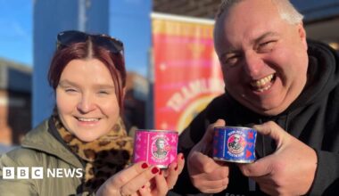 A woman with red hair wearing a thick green winter coat stands next to a jolly-looking man in a black hoodie. They are both holding up miniature cans of beans featuring pictures of Rick Astley and Fatboy Slim.