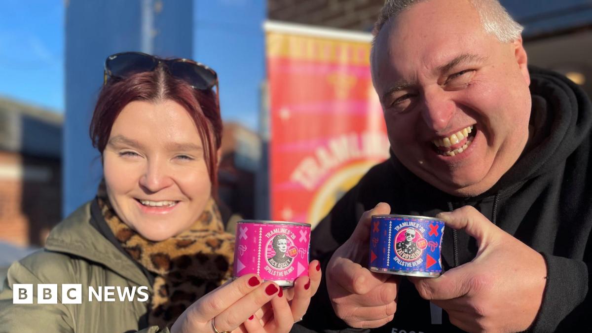 A woman with red hair wearing a thick green winter coat stands next to a jolly-looking man in a black hoodie. They are both holding up miniature cans of beans featuring pictures of Rick Astley and Fatboy Slim.