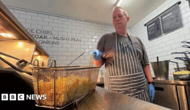 Paul Costelloe has receding grey hair. He is wearing a bright pink, blue and green scarf and a blue jacket.